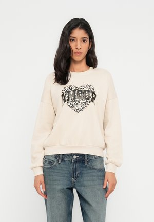 Beige sweatshirt met lange mouwen, geribbelde zoom en een hartvormig luipaardprint ontwerp met het woord "AMOUR" in vetgedrukte letters. Draagt over een blauwe spijkerbroek.