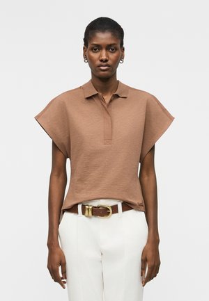 Femme portant un haut marron à manches courtes avec col, un pantalon blanc et une ceinture marron avec boucle dorée, se tenant devant un fond uni.