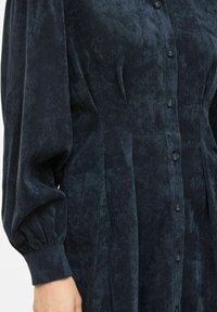 Teksturirana tamnoplava košulja-haljina od korduroga s gumbima sprijeda, naboranim rukavima i bočnim džepovima, ističući uzorak rebraste tkanine.