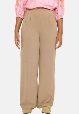 Kobieta ubrana w beżowe spodnie o szerokich nogawkach, połączone z jasnobrązowymi mokasynami oraz różową bluzką w paski z haftowanymi rękawami.