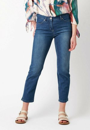 Woman wearing blue cropped jeans, a colorful abstract blouse, and beige open-toe sandals, standing against a white background.