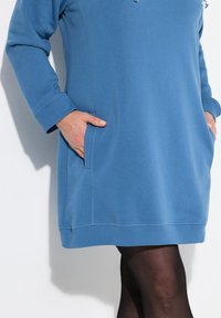 Blaues Sweatkleid mit langen Ärmeln, seitlichen Taschen und gerippten Bündchen. Es hat eine glatte Textur und eine lockere Passform und wird über einer schwarzen Strumpfhose getragen.
