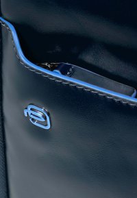 Bolso de cuero marino con textura suave, que presenta un acento azul claro a lo largo del borde y un emblema de logo metálico en el costado.