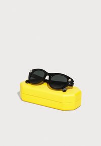 Ochelari de soare negri cu lentile ovale, având un cadru din plastic neted și un accent de cristale transparente, așezați pe un toc texturat galben.