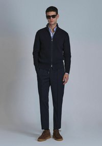 Marineblaue gerippte Zip-Up-Strickjacke, darunter ein hellblaues Hemd mit Kragen, kombiniert mit dunkel maßgeschneiderten Hosen und braunen Wildlederschuhen mit Gummisohlen.