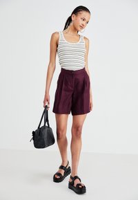 Stripete singlet, høytlivede burgunder-shorts, svarte platåsandaler og en svart lærveske, satt mot en ensfarget bakgrunn.