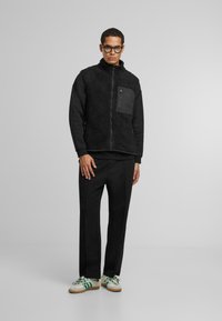 Schwarze Fleece-Weste mit einer Reißverschlusstasche, über einem schwarzen Langarmshirt getragen. Kombiniert mit schwarzen Hosen und weißen Sneakern mit grünen Akzenten.