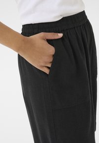 Pantaloni neri, larghi e comodi, con vita elastica e tasche laterali, realizzati in tessuto testurizzato, mostrati con una mano nella tasca.