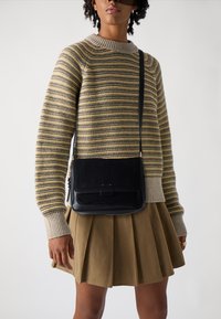 Borsa a tracolla nera con un mix di camoscio e pelle, dotata di chiusura a pattina, tasca con zip e tracolla regolabile. Indossata con un maglione a righe e una gonna.