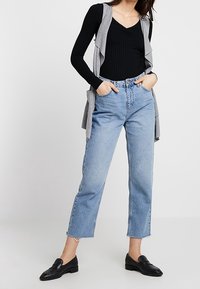 Szara bezrękawnik na czarnym prążkowanym topie, w zestawieniu z jasnoniebieskimi prostymi dżinsami i czarnymi loafersami. Luźny strój z warstwowymi fakturami.