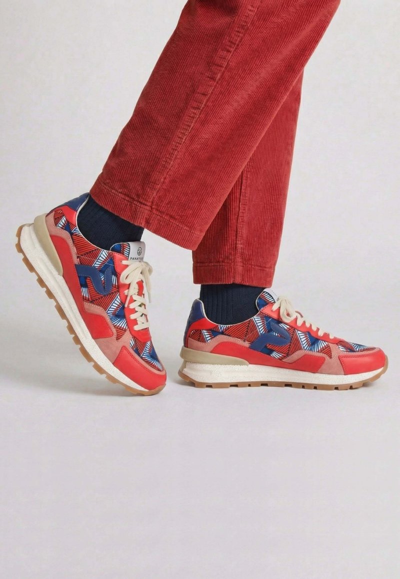 Pieds portant des baskets rouges et bleues à motifs avec des lacets blancs, des chaussettes noires et un pantalon en velours côtelé rouge sur un fond clair uni.