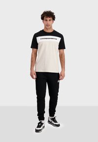 T-shirt con maniche nere e corpo bianco, con un logo nero; abbinato a pantaloni della tuta neri e sneakers bianco e nere.