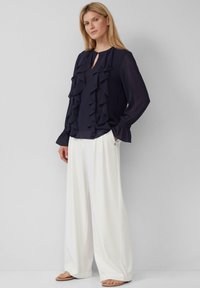 Blusa blu navy con volants sovrapposti, apertura a chiave sul colletto e maniche lunghe; abbinata a pantaloni bianchi a gamba larga e sandali.