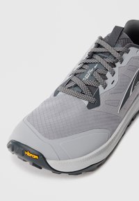 Baskets de sport grises et noires avec des lacets à motifs, semelle Vibram et tige en mesh respirant, inclinées pour montrer les détails avant et latéral.