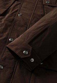 Mørkebrune jakkeærmer med sytter, metal trykknapper og en, der er graveret med et mærkenavn.