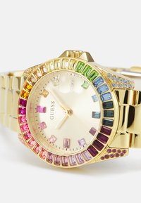 Montre dorée avec un cadran orné de pierres précieuses multicolores en rectangles et ronds, un cadran argenté et un bracelet en maillons.