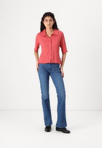Chemise à boutons rouge rayée avec des manches courtes, associée à un jean bleu évasé. La chemise présente un ourlet festonné et un tissu texturé.