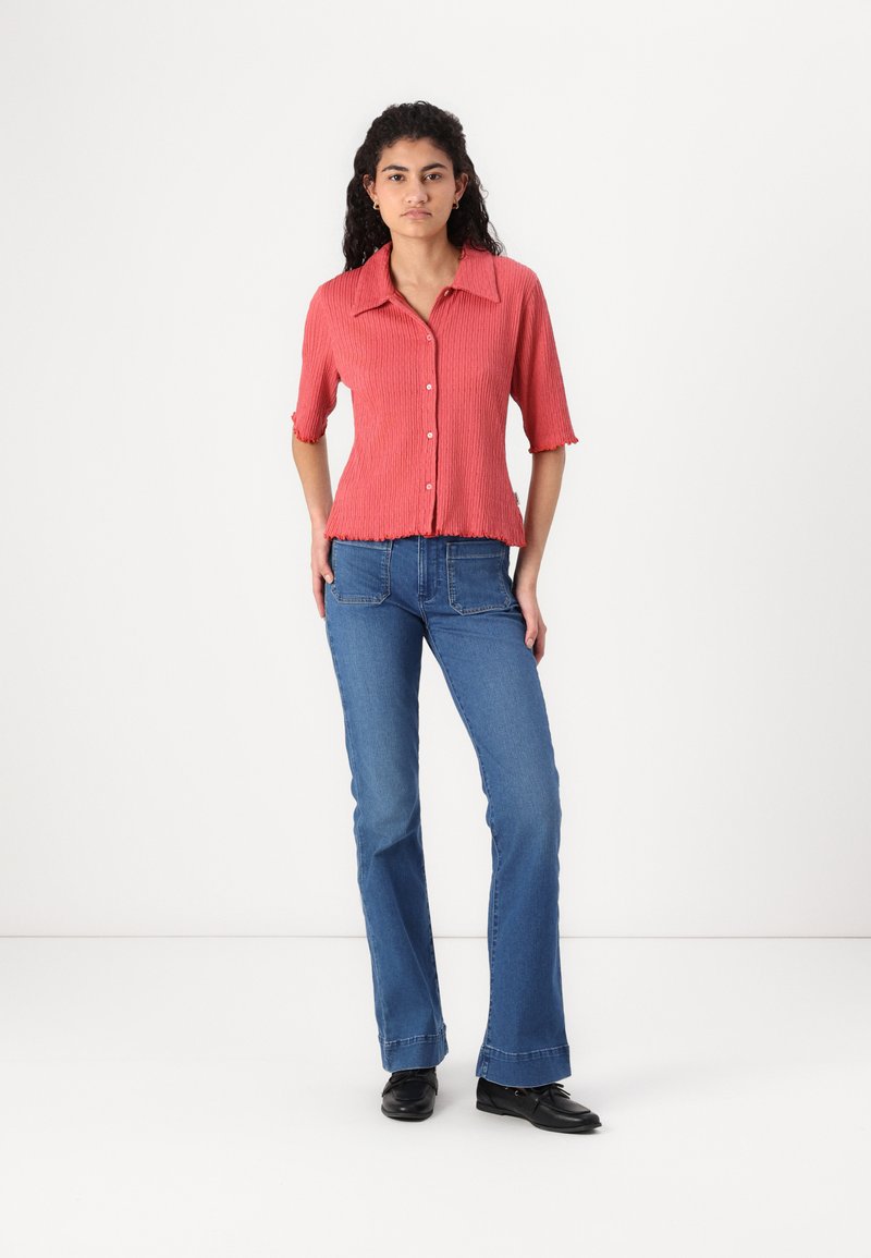 Camicia rossa a righe con bottoni e maniche corte, abbinata a jeans a zampa d'elefante blu. La camicia presenta un orlo ondulato e un tessuto testurizzato.