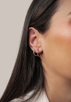 Nahaufnahme des linken Ohrs einer Person mit zwei kleinen silbernen Creolen und langen dunklen Haaren, die hinter das Ohr gesteckt sind.