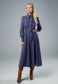 Długa niebieska sukienka z kołnierzykiem, wzorzystą teksturą, zapięciem na guziki, cinchowanym wcięciem w talii oraz brązowym paskiem; w połączeniu z brązowymi botkami do kolan.