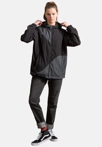 Zwarte en grijze windbreaker jas met capuchon, voorzien van een geometrisch ontwerp, ritssluiting en elastische manchetten, gecombineerd met donkere jeans en zwarte sneakers.