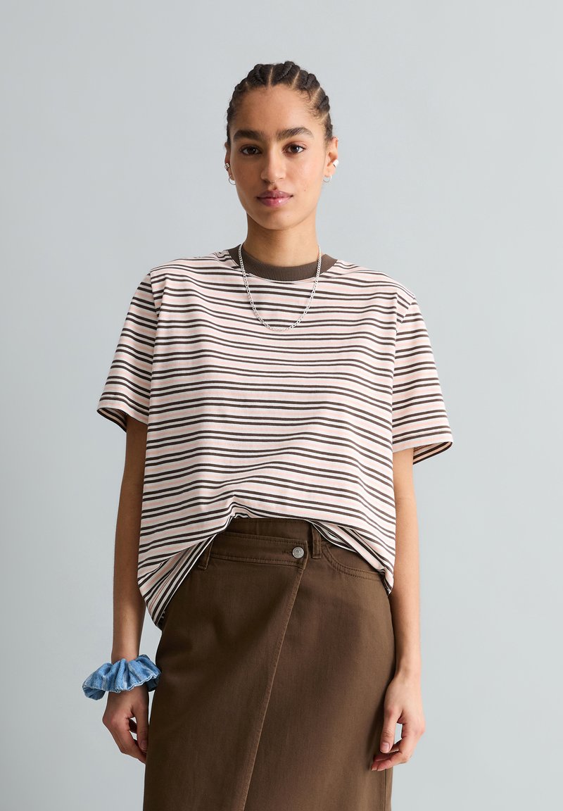 Jeune femme aux cheveux tressés portant un t-shirt rayé rentré dans une jupe marron, avec un chouchou bleu au poignet.