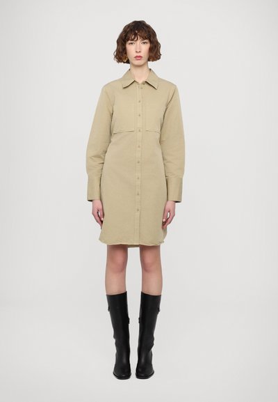 Vestido camisas color caqui con mangas largas, frente de botones y un bolsillo en el pecho, combinado con botas negras hasta la rodilla. Textura de tela suave.
