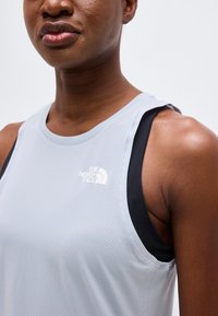 Κοντινό πλάνο ενός ατόμου που φοράει αμάνικη αθλητική μπλούζα ανοιχτό γκρι με μαύρο αθλητικό σουτιέν από κάτω, με το λογότυπο "The North Face" εμφανές.