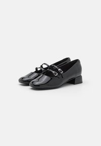 Zapatos de charol negro con un tacón bajo y cuadrado. Presentan dos correas ajustables con hebillas y un diseño de punta redondeada.