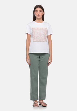 Giovane donna in piedi che indossa una camicia bianca a maniche corte con un disegno floreale quadrato rosa, pantaloni verdi e sandali bianchi con cinturini.