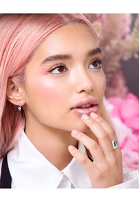 Close-up of a person with pink hair, wearing silver earrings and a wide silver ring, with glossy lips and subtle eye makeup.