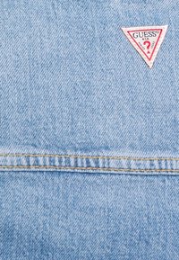 Světlá modrá džínovina s malou červenobílou trojúhelníkovou nášivkou s logem "GUESS" a otazníkem. Viditelné detailní šití.