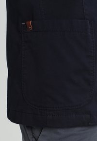 Granatowa kurtka z dużą przednią kieszenią z brązowym skórzanym akcentem, płaskimi szwami oraz miękką teksturą bawełnianego materiału.