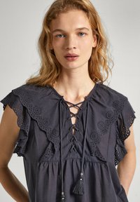 Blusa grigia scura con volant in pizzo ad occhio, apertura frontale con lacci e dettagli con frange. Tessuto morbido con una vestibilità ampia, caratterizzata da un collo rotondo.