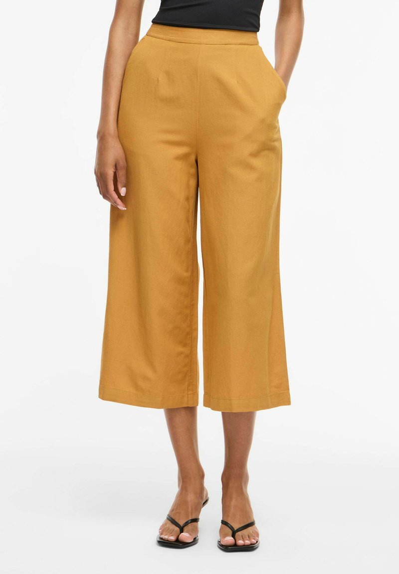 Pantalon large moutarde taille haute, court, porté avec un top noir sans manches et des sandales noires à talons et à lanières. Mains dans les poches.