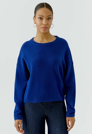Vrouw met donker haar naar achteren gebonden, draagt oorbellen in de vorm van ringen, een blauwe trui met lange mouwen en donkere jeans, staande tegen een effen achtergrond.