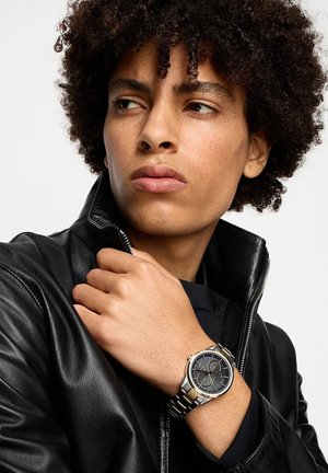 Mlad človek s kodrastimi lasmi, oblečen v črno usnjeno jakno, prilagaja ovratnik in prikazuje srebrno-zlato uro z black face.