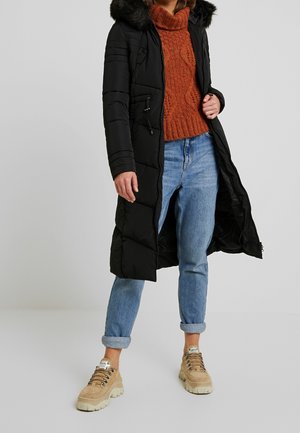 Personne portant un long manteau matelassé noir avec capuche en fourrure, un pull en maille orange rouille, un jean bleu retroussé et des baskets beiges épaisses.