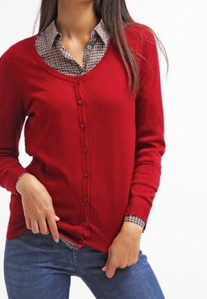 Femme portant un cardigan rouge boutonné sur une chemise à motifs avec un jean bleu, montrée des épaules aux hanches.