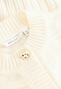 Cremefarbener, gestrickter Cardigan mit gerippter Struktur. Verfügt über einen runden Knopf in Goldoptik und ein Etikett mit Größen- und Markenangabe, Noella.