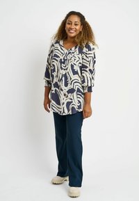 Blusa blu navy ampia con motivo astratto crema, maniche a tre quarti e scollatura tonda, abbinata a pantaloni scuri e scarpe chiare.