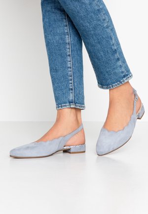 Osoba nosząca jasnoniebieskie baleriny typu slingback z falistym wykończeniem, niskim obcasem oraz podwinięte niebieskie dżinsy na białej podłodze.