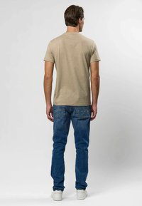 Mees seisab seljaga vaataja poole, seljas beež lühikeste varrukatega särk, sinised teksad ja valged tossud, taustaks hele ühtlane pind.