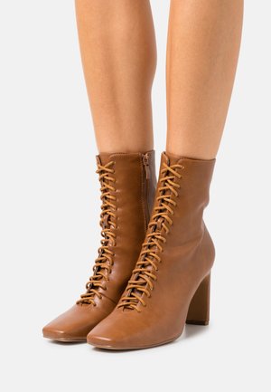 Læderankelstøvler i tan med snøre og blokhæle båret på bare ben mod en ensfarvet hvid baggrund.