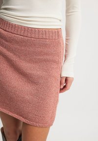 Pletena svinčnik krila v koralni barvi z teksturiranim zaključkom. Ima rebrast pas in ravno krojenje, ki sega do sredine stegna.