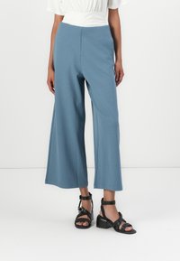 Lichtblauwe wijde broek van zacht materiaal met een gladde textuur, gecombineerd met zwarte haksandalen en een witte top.