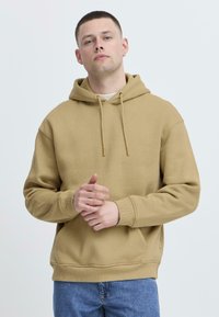 Tan hættetrøje lavet af blødt stof med snoretræk, kengurulomme og ribbede manchetter; parret med blå jeans.