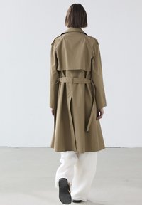 Khakifarbener Trenchcoat mit einem Gürtel, seitlichen Schlitzen und einem geschichteten Rücken-Design. Über weißen Hosen getragen, betont er seinen maßgeschneiderten Look und die glatte Textur.