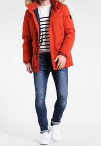 Červená zimní bunda s polstrováním a kapucí lemovanou kožešinou, tmavě modrý vnitřek, zapínání na knoflíky a několik kapes, kombinovaná s modrými džínami a bílými teniskami.