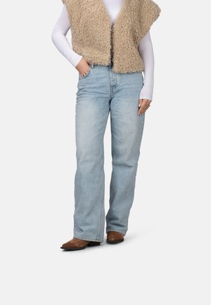 Gilet in pelliccia sintetica beige sopra una camicia bianca a maniche lunghe, abbinato a jeans azzurri larghi e stivali alla caviglia marroni con tacco.